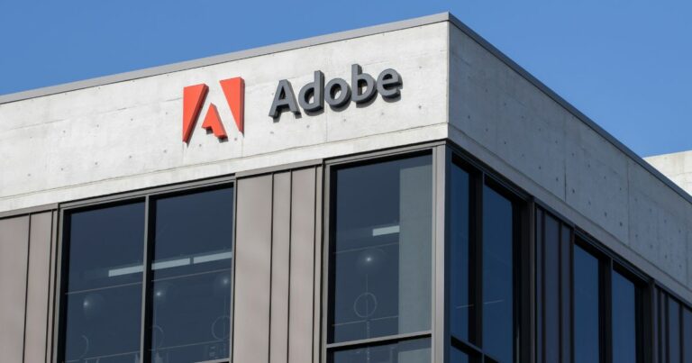 Adobe, marketing, Fachada de um prédio corporativo moderno com o logotipo da Adobe no topo, exibindo o símbolo vermelho e o nome da empresa em letras pretas sobre uma estrutura de concreto claro e janelas amplas de vidro, sob um céu azul