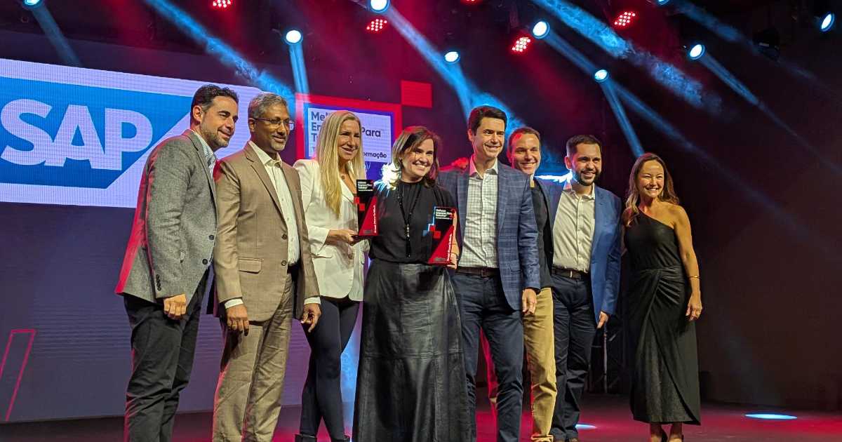 Grupo de oito pessoas em um palco de premiação, posando para a foto sob luzes coloridas. Ao centro, duas mulheres seguram troféus, enquanto demais participantes sorriem ao lado delas. Ao fundo, aparece um telão iluminado com o logo da SAP e a referência ao prêmio GPTW, empresas