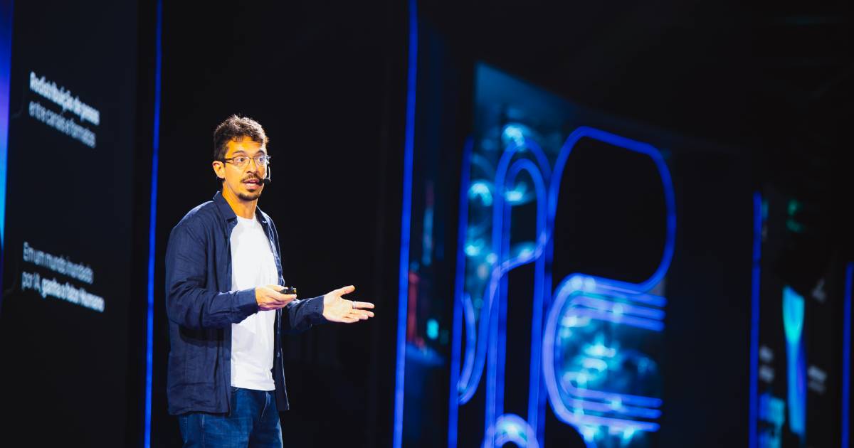 Gustavo Avelar, vice-presidente da RD Station, fala em um palco iluminado com luzes azuis durante o RD Summit, segurando um controle e utilizando microfone de cabeça.