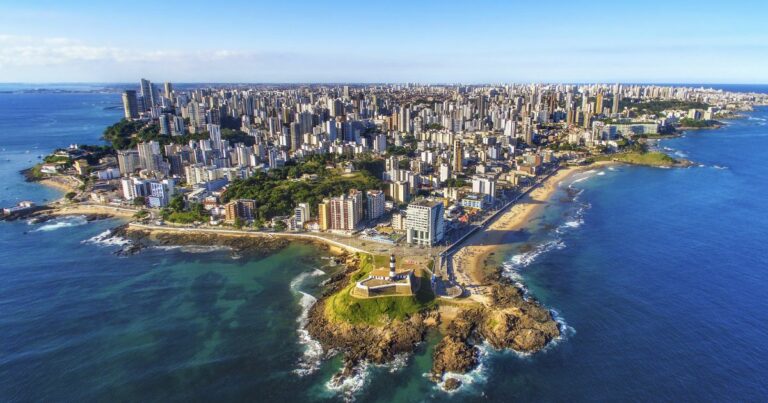 A foto mostra uma vista aérea de uma cidade litorânea com uma extensa faixa de prédios altos ao fundo e várias praias ao longo da costa. Em primeiro plano, há uma pequena península com um farol histórico e construções ao redor, cercada por águas azuis e áreas rochosas. O mar é amplo e ocupa grande parte da imagem, com tons que variam do verde próximo à costa ao azul profundo mais distante. O céu está claro, indicando um dia ensolarado. (futuro)