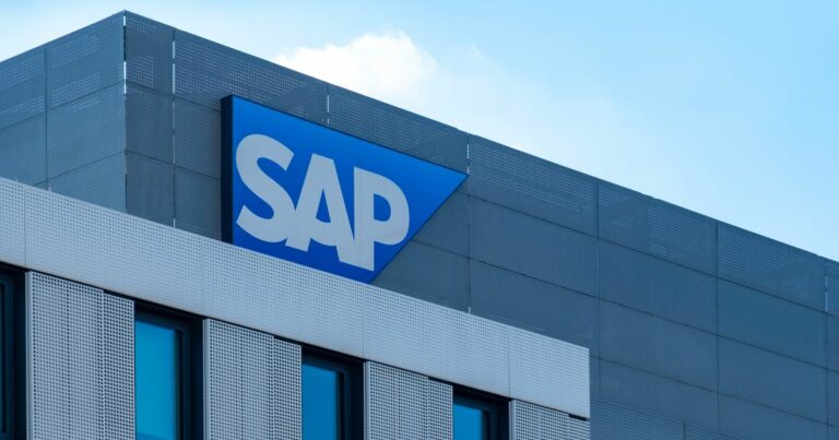 A foto mostra a fachada de um edifício corporativo com o logotipo da SAP em destaque. O logotipo está em letras brancas sobre um fundo azul triangular, posicionado na parte superior da estrutura. O prédio tem revestimento metálico cinza com painéis perfurados e janelas de vidro azuladas. O céu ao fundo está claro, sugerindo um dia ensolarado, graduação