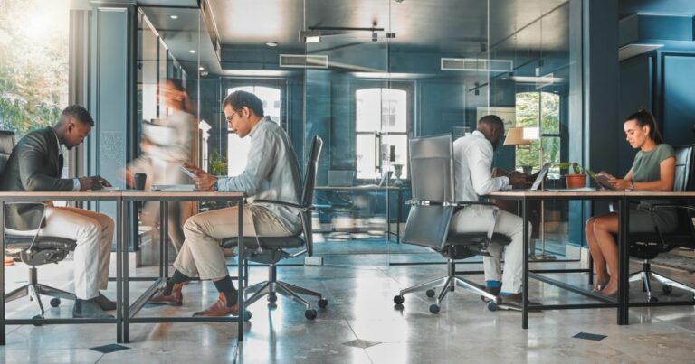 A foto mostra um ambiente corporativo moderno com mesas compartilhadas e cadeiras ergonômicas. Há quatro pessoas sentadas trabalhando em laptops e dispositivos móveis, enquanto outra pessoa caminha ao fundo, criando uma sensação de movimento. O espaço é iluminado por luz natural que entra pelas janelas, e as paredes são de vidro, separando áreas internas. O piso é claro e polido, e há elementos decorativos discretos, como plantas sobre as mesas. (tecnologia)