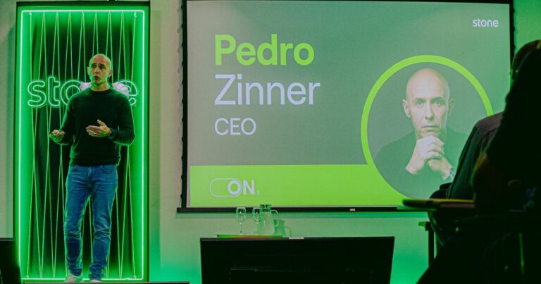 A imagem mostra um evento corporativo com identidade visual predominante em tons de verde, associado à marca Stone. No palco, há uma pessoa apresentando, posicionada à esquerda, diante de um painel iluminado com linhas verticais e o logotipo da empresa. À direita, um grande telão exibe informações do palestrante: Texto visível: “Pedro Zinner” em destaque, com “CEO” logo abaixo. Logotipo da Stone no canto superior direito. Elemento gráfico circular com a foto do palestrante. Palavra “ON.” na parte inferior do slide. No primeiro plano, há uma mesa com copos e garrafas de água, sugerindo um ambiente preparado para uma apresentação formal. A iluminação é intensa, com foco no verde, reforçando a identidade da marca e criando um clima moderno e tecnológico.