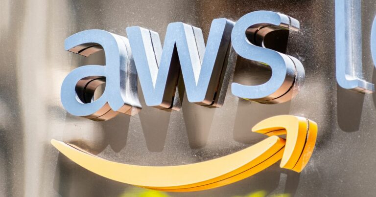 A imagem mostra o logotipo da AWS (Amazon Web Services) em destaque, com letras tridimensionais em tom azul sobre um fundo metálico. Abaixo do texto, aparece a clássica seta curva da Amazon em cor amarela, reforçando a identidade visual da marca. O design é limpo e moderno, com iluminação que cria reflexos sutis no fundo, transmitindo sofisticação e tecnologia. Esse tipo de composição é típico de sinalização corporativa ou branding em eventos e escritórios.