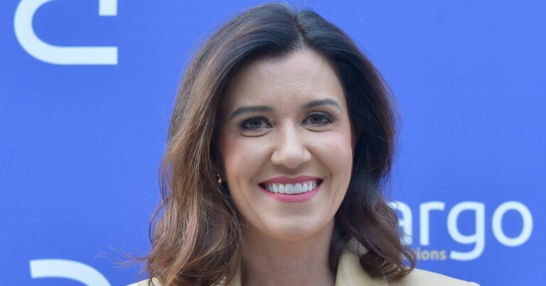 A imagem mostra uma pessoa com cabelos castanhos, ondulados e comprimento médio, vestindo um blazer claro. O fundo é azul, com parte de um logotipo visível à direita, contendo as letras “argo” e um texto menor abaixo, sugerindo que seja um painel ou backdrop de evento corporativo ou conferência.