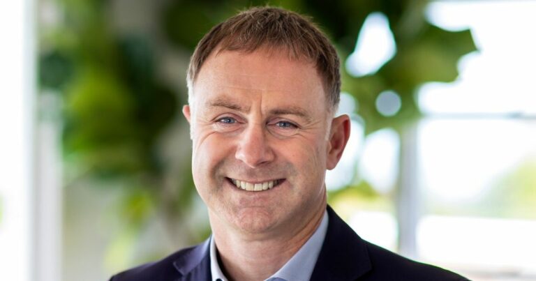 A imagem mostra uma pessoa em um ambiente corporativo ou profissional, com fundo desfocado que parece ser um espaço interno iluminado por luz natural. (Nova Zelândia)