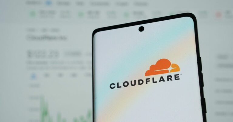A imagem mostra um smartphone em primeiro plano exibindo o logotipo da Cloudflare, uma empresa conhecida por serviços de segurança, desempenho e confiabilidade na internet. O fundo está desfocado, mas parece mostrar uma tela com gráficos e números, possivelmente relacionados a dados financeiros ou estatísticas.