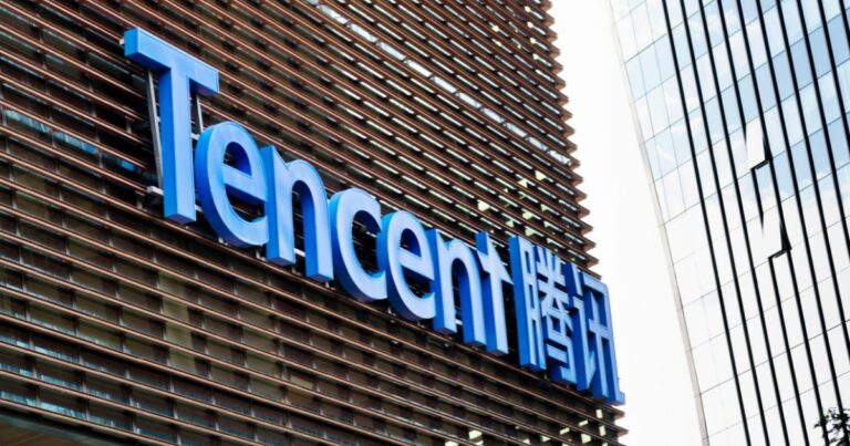 A foto mostra a fachada de um edifício corporativo com o logotipo da Tencent em destaque. O nome da empresa aparece em letras grandes azuis, acompanhado por caracteres chineses à direita, também em azul. A estrutura do prédio tem painéis horizontais metálicos e, ao fundo, é possível ver parte de outro edifício com fachada de vidro espelhado. O céu está claro, sugerindo um dia ensolarado.