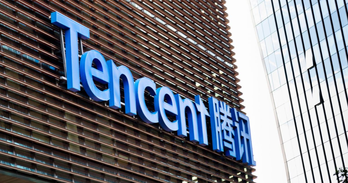 A foto mostra a fachada de um edifício corporativo com o logotipo da Tencent em destaque. O nome da empresa aparece em letras grandes azuis, acompanhado por caracteres chineses à direita, também em azul. A estrutura do prédio tem painéis horizontais metálicos e, ao fundo, é possível ver parte de outro edifício com fachada de vidro espelhado. O céu está claro, sugerindo um dia ensolarado.