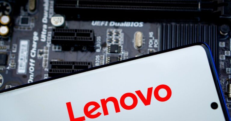 A foto mostra um dispositivo eletrônico com a marca Lenovo em destaque, escrita em letras vermelhas sobre fundo branco, provavelmente na tela de um smartphone ou tablet. Ao fundo, desfocado, há uma placa-mãe com componentes visíveis, incluindo slots de expansão e texto técnico como “UEFI DualBIOS”. A imagem transmite um contexto tecnológico, associando a marca Lenovo a hardware e sistemas.
