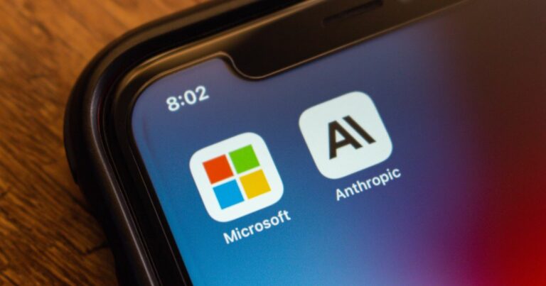 A foto mostra a tela de um smartphone com dois ícones de aplicativos em destaque: Microsoft, com o logotipo colorido em forma de quadrados, e Anthropic, com um ícone minimalista contendo as letras “AI”. O fundo da tela tem um gradiente em tons de azul e roxo, e no canto superior esquerdo aparece o horário 8:02. A borda do aparelho é visível, apoiada sobre uma superfície de madeira.