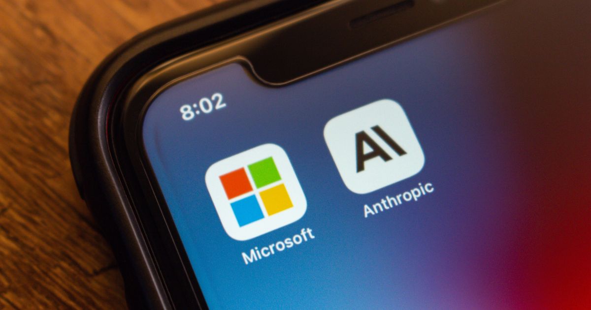 A foto mostra a tela de um smartphone com dois ícones de aplicativos em destaque: Microsoft, com o logotipo colorido em forma de quadrados, e Anthropic, com um ícone minimalista contendo as letras “AI”. O fundo da tela tem um gradiente em tons de azul e roxo, e no canto superior esquerdo aparece o horário 8:02. A borda do aparelho é visível, apoiada sobre uma superfície de madeira.