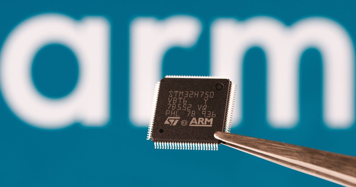 A imagem mostra um chip eletrônico sendo segurado por uma pinça metálica, com um fundo azul que exibe a palavra arm em letras grandes e brancas, indicando relação com a empresa ARM, conhecida por projetar arquiteturas de processadores.