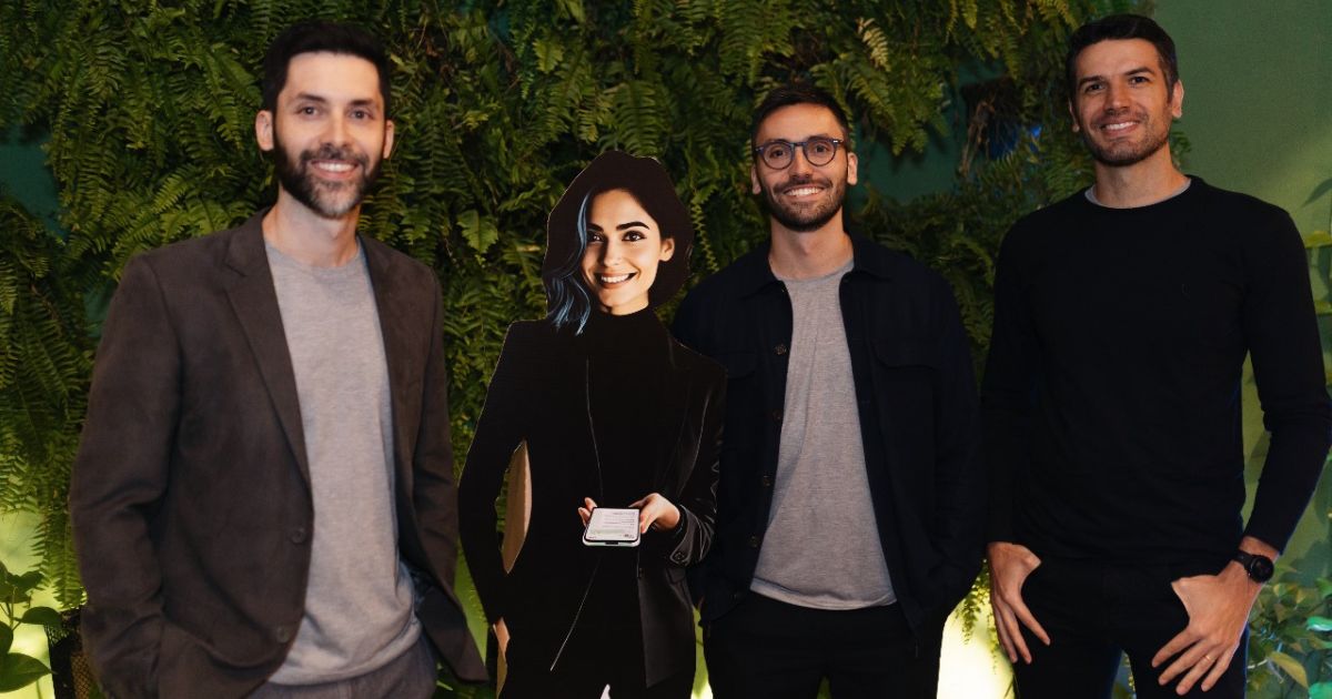 A imagem mostra três pessoas em pé, posicionadas diante de um fundo verde com folhagens, sugerindo um ambiente natural ou decorado para um evento. Elementos visuais principais: As pessoas estão vestindo roupas formais e casuais: uma com terno cinza e camiseta clara, outra com camisa escura sobre camiseta cinza, e a terceira com camiseta preta e calça escura. No centro, há uma silhueta recortada de papelão ou material similar, representando uma figura humana segurando um smartphone, o que indica um elemento decorativo ou promocional. Contexto: A composição sugere um encontro corporativo, networking ou evento social com identidade visual moderna e descontraída, reforçada pelo cenário verde e iluminação suave. (Agendor)