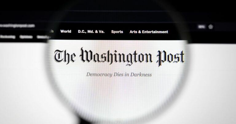 A foto mostra uma tela com o site do jornal The Washington Post ampliado por uma lente de aumento. O logotipo do jornal aparece em destaque na parte superior, com a frase “Democracy Dies in Darkness” logo abaixo. Na barra de navegação superior, são visíveis seções como “World”, “D.C., Md. & Va.”, “Sports” e “Arts & Entertainment”. O fundo é predominantemente branco, com detalhes em preto, e a lente cria um efeito de foco sobre o nome do jornal. (Oracle)
