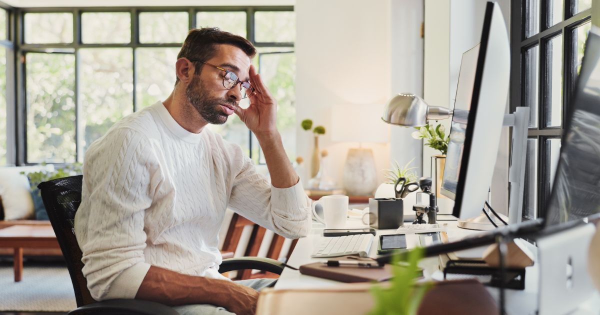 A foto mostra uma pessoa sentada em uma mesa de trabalho moderna, em frente a um computador com tela grande. Sobre a mesa há teclado, mouse, caneca, caderno, óculos e outros objetos organizados, além de uma luminária metálica e um vaso com flores. Ao fundo, há uma sala iluminada por luz natural que entra por amplas janelas, com móveis como sofá e mesa lateral, criando um ambiente aconchegante e bem decorado. (CISO)