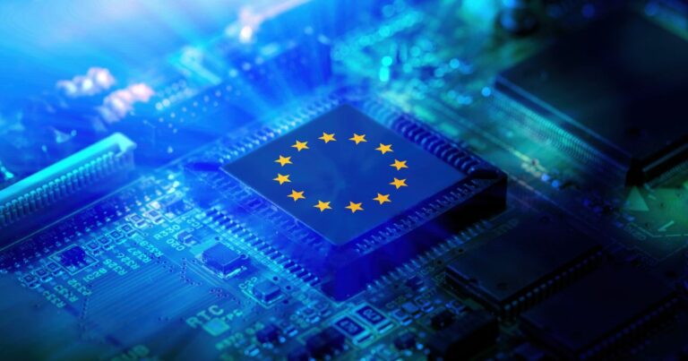 A foto mostra um close-up de uma placa de circuito eletrônico iluminada por luz azul intensa. No centro, há um chip com a bandeira da União Europeia estampada, composta por um círculo de estrelas amarelas sobre fundo azul. A imagem transmite uma ideia de tecnologia e integração europeia, sugerindo temas como inovação, indústria de semicondutores ou políticas digitais.