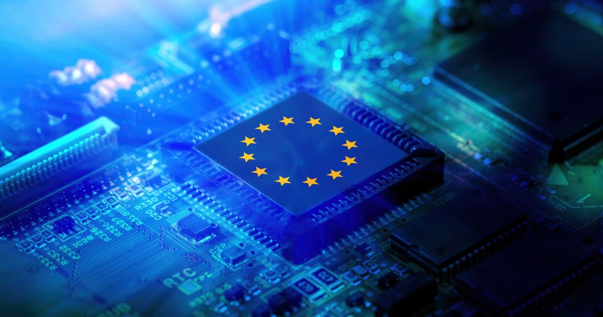 A foto mostra um close-up de uma placa de circuito eletrônico iluminada por luz azul intensa. No centro, há um chip com a bandeira da União Europeia estampada, composta por um círculo de estrelas amarelas sobre fundo azul. A imagem transmite uma ideia de tecnologia e integração europeia, sugerindo temas como inovação, indústria de semicondutores ou políticas digitais.