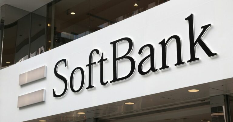 A foto mostra a fachada de um edifício com o logotipo da SoftBank em destaque. O nome da empresa aparece em letras pretas grandes sobre um fundo branco, acompanhado por dois retângulos horizontais cinza à esquerda, que fazem parte da marca. A estrutura é moderna, com linhas limpas e iluminação suave, sugerindo um ambiente corporativo.