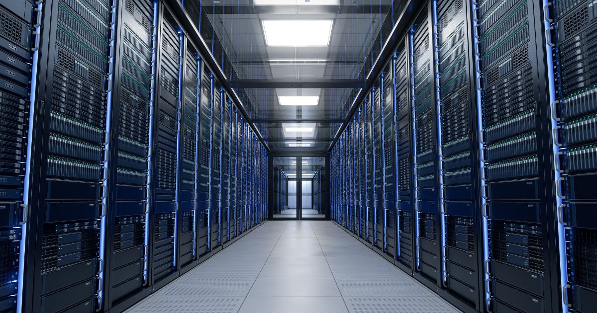 A foto mostra o interior de um corredor de data center, com fileiras de racks de servidores alinhadas dos dois lados. Cada rack contém múltiplos equipamentos eletrônicos organizados verticalmente, com luzes indicadoras verdes e azuis acesas. O ambiente é iluminado por painéis de luz no teto, refletindo no piso claro e criando uma aparência limpa e moderna. A perspectiva central dá profundidade à imagem, destacando a simetria e a infraestrutura tecnológica. (IEA), computação