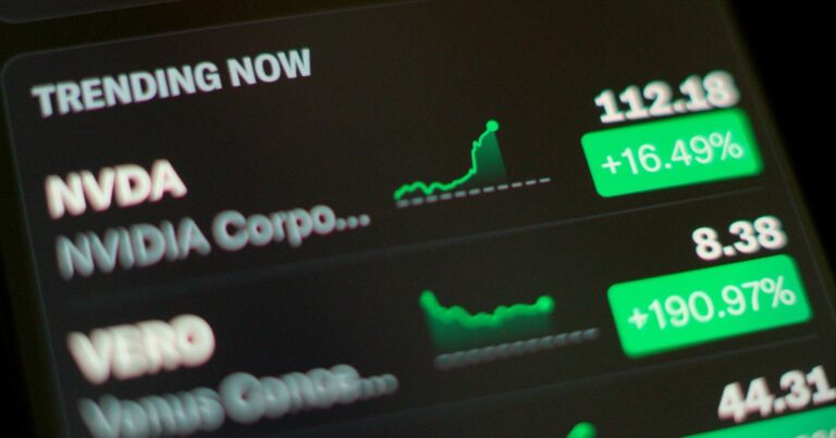 A foto mostra uma tela com informações financeiras, destacando ações em alta. No topo, aparece a seção “TRENDING NOW” em letras brancas sobre fundo preto. Logo abaixo, há dados da empresa NVIDIA (símbolo NVDA), com preço de 112,18 e variação positiva de +16,49%, acompanhada por um gráfico de linha ascendente em verde. Em seguida, há informações da empresa VERO, com preço de 8,38 e variação expressiva de +190,97%, também com gráfico verde. A interface tem um design escuro, com números e indicadores em verde para ganhos. (tecnologia)