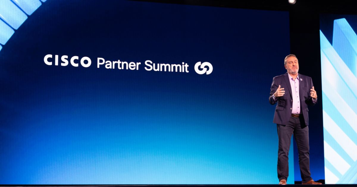 A cena mostra uma pessoa em pé sobre um palco durante um evento corporativo. No fundo, há uma grande tela azul com o texto “CISCO Partner Summit” acompanhado de um ícone gráfico. A pessoa está vestindo um traje formal composto por paletó escuro, camisa clara e calça social, e parece estar fazendo uma apresentação ou discurso, com gestos que indicam comunicação ativa. A iluminação é profissional, destacando o apresentador e criando um ambiente típico de conferência empresarial.