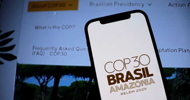A cena mostra um smartphone em primeiro plano exibindo o logotipo e texto “COP30 BRASIL AMAZÔNIA BELÉM 2025” em letras marrons sobre fundo claro. Ao fundo, desfocado, há uma tela com informações sobre a COP30, incluindo seções como “About COP30”, “Brazilian Presidency”, e perguntas frequentes (FAQ – COP30). A composição sugere um contexto relacionado à conferência climática COP30, destacando sua realização no Brasil, na Amazônia, em Belém, no ano de 2025.