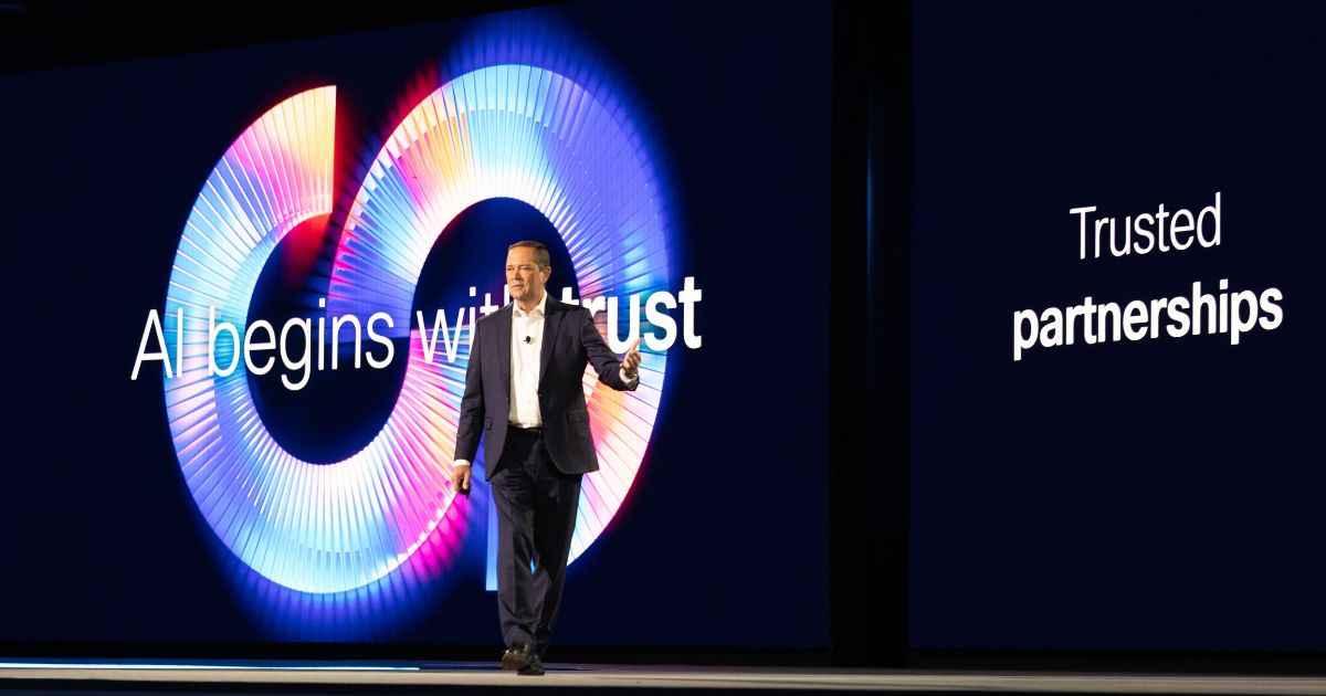 A cena mostra uma pessoa em pé sobre um palco, vestindo traje formal com paletó escuro, camisa clara e sapatos sociais. Ao fundo, há uma grande tela com design moderno em tons de azul, roxo e rosa, exibindo um círculo estilizado com efeito de luz. O texto visível na tela diz: “AI begins with trust” e, à direita, “Trusted partnerships”. A iluminação é intensa e direcionada para destacar o apresentador, criando um ambiente típico de conferência tecnológica ou evento corporativo de grande porte. (Cisco)