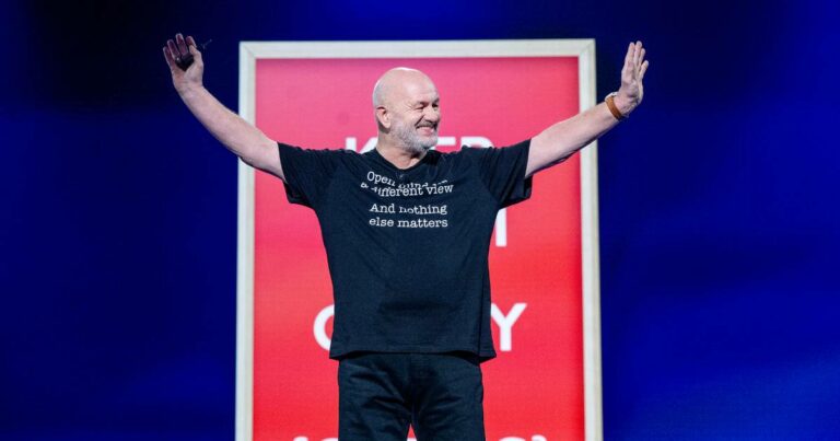 Homem calvo identificado como Werner Vogels, vice-presidente e CTO da Amazon, em pé no palco de um evento, sorrindo com os braços abertos; ele usa camiseta preta com frase em inglês e há um painel vermelho desfocado ao fundo