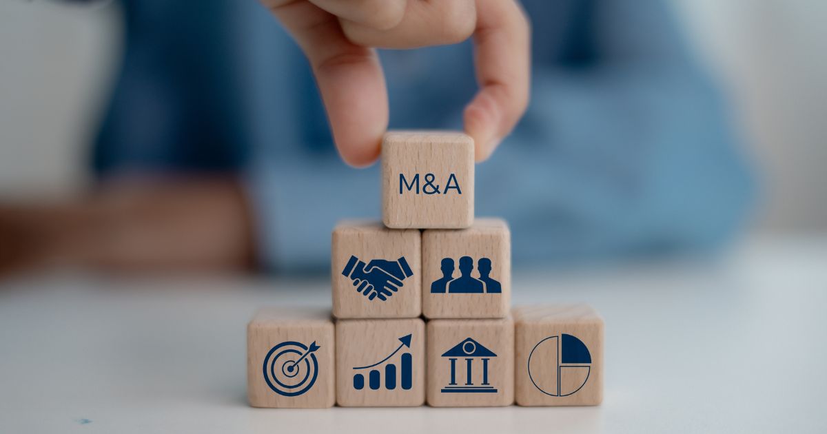 A imagem mostra uma mão posicionando um bloco de madeira no topo de uma estrutura formada por outros blocos, criando uma pirâmide. O bloco superior tem a inscrição “M&A”, que se refere a fusões e aquisições (Mergers and Acquisitions). Os blocos da base apresentam ícones relacionados a negócios e finanças: um alvo com uma flecha, um gráfico de crescimento, um edifício com colunas (representando instituições financeiras), um gráfico de pizza e, na segunda camada, ícones de aperto de mãos (parceria) e um grupo de pessoas (equipe ou stakeholders). O fundo é desfocado, com tons de azul, sugerindo um ambiente corporativo ou profissional.