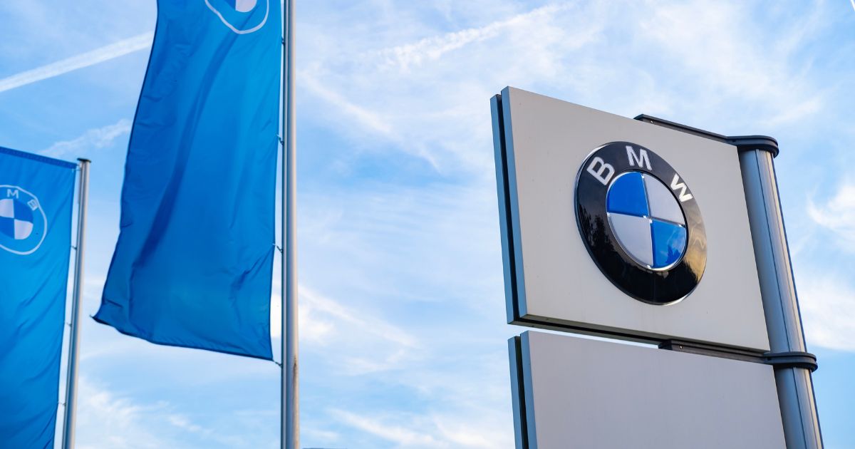 A imagem mostra um cenário externo com elementos relacionados à marca BMW. Aqui estão os detalhes principais: Contexto geral: Céu azul com algumas nuvens finas e trilhas de avião, sugerindo um dia claro e ensolarado. Elementos visíveis: Duas bandeiras verticais azuis, cada uma com o logotipo da BMW em branco, posicionadas à esquerda. Um painel ou placa vertical à direita, com o logotipo da BMW em destaque (círculo dividido em quadrantes azul e branco, com a sigla “BMW” na parte superior). Cores predominantes: Azul (bandeiras e céu), branco (detalhes do logotipo e fundo da placa), e preto (bordas do logotipo). Atmosfera: Limpa, corporativa e moderna, transmitindo identidade visual forte da marca. Esse tipo de imagem é ideal para materiais institucionais, posts sobre automóveis premium, ou conteúdos relacionados à inovação e tradição da BMW.