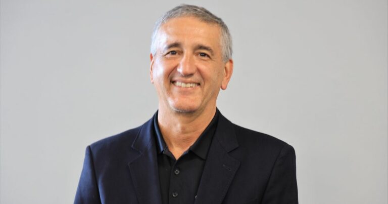 A imagem mostra uma pessoa vestindo um blazer escuro sobre uma camisa polo preta, com botões visíveis. O fundo é liso e claro, provavelmente cinza, sem elementos adicionais ou distrações, sugerindo um ambiente neutro, como um estúdio ou espaço corporativo. A iluminação é uniforme, destacando a roupa e a postura da pessoa, que está posicionada de forma centralizada na imagem. (Stefanini)