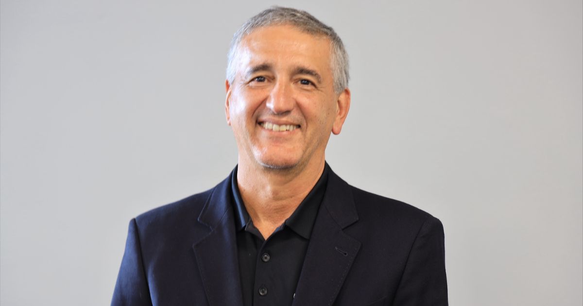 A imagem mostra uma pessoa vestindo um blazer escuro sobre uma camisa polo preta, com botões visíveis. O fundo é liso e claro, provavelmente cinza, sem elementos adicionais ou distrações, sugerindo um ambiente neutro, como um estúdio ou espaço corporativo. A iluminação é uniforme, destacando a roupa e a postura da pessoa, que está posicionada de forma centralizada na imagem. (Stefanini)