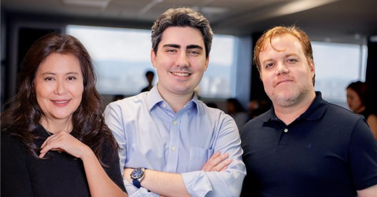 A imagem mostra três pessoas posicionadas lado a lado em um ambiente corporativo interno. A pessoa ao centro veste uma camisa social azul clara com mangas dobradas e um relógio no pulso esquerdo, com os braços cruzados. À esquerda, a pessoa veste uma blusa preta de manga longa, com uma das mãos apoiada no braço oposto. À direita, a pessoa usa uma camisa polo preta. O fundo apresenta um espaço de escritório com iluminação artificial, janelas ao fundo que revelam luz natural e parte de uma paisagem externa. Há outras pessoas desfocadas ao fundo, sugerindo um ambiente de reunião ou evento corporativo. (Numen)