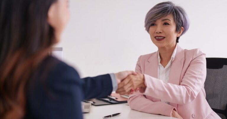 Duas pessoas estão sentadas frente a frente em um ambiente corporativo claro e organizado. Elas estão apertando as mãos sobre uma mesa branca, sugerindo um acordo, entrevista ou reunião profissional. A pessoa à direita veste um blazer rosa claro sobre uma camisa branca, enquanto a pessoa à esquerda usa um blazer escuro. Sobre a mesa há alguns itens de trabalho, como papéis, uma caneta e um teclado, compondo um cenário de escritório moderno. (ano)