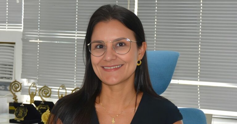 A imagem mostra uma pessoa sentada em uma cadeira de escritório azul, usando uma blusa preta e acessórios dourados — incluindo brincos circulares e um colar delicado. Ao fundo, há uma parede com persianas cinza fechadas e uma mesa onde estão dispostos vários troféus dourados, sugerindo um ambiente corporativo ou uma sala de premiações. A iluminação é neutra e destaca os elementos do cenário. (justiça)