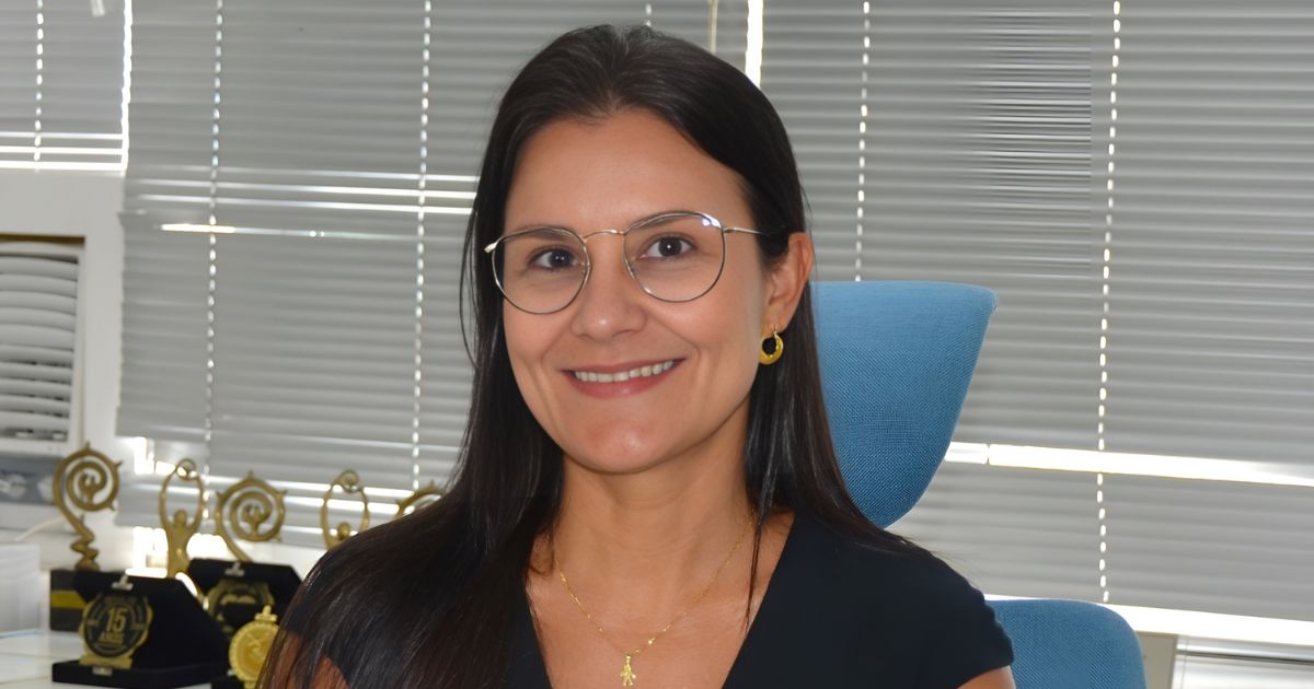 A imagem mostra uma pessoa sentada em uma cadeira de escritório azul, usando uma blusa preta e acessórios dourados — incluindo brincos circulares e um colar delicado. Ao fundo, há uma parede com persianas cinza fechadas e uma mesa onde estão dispostos vários troféus dourados, sugerindo um ambiente corporativo ou uma sala de premiações. A iluminação é neutra e destaca os elementos do cenário. (justiça)