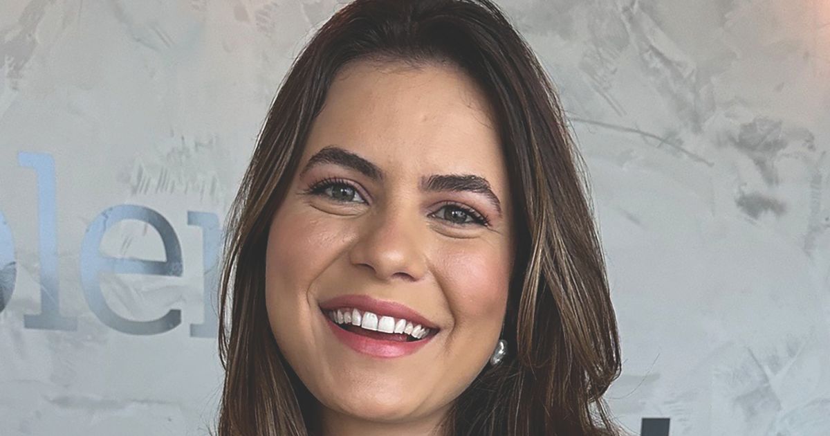 A imagem mostra uma pessoa de cabelos castanhos com mechas mais claras, soltos e levemente ondulados, posicionada em frente a uma parede texturizada em tons de cinza. Ao fundo, é possível ver parte de um texto aplicado à parede, com letras grandes e metálicas parcialmente visíveis. A iluminação suave destaca tanto a textura do fundo quanto os fios de cabelo, criando um ambiente moderno e minimalista. (IA)