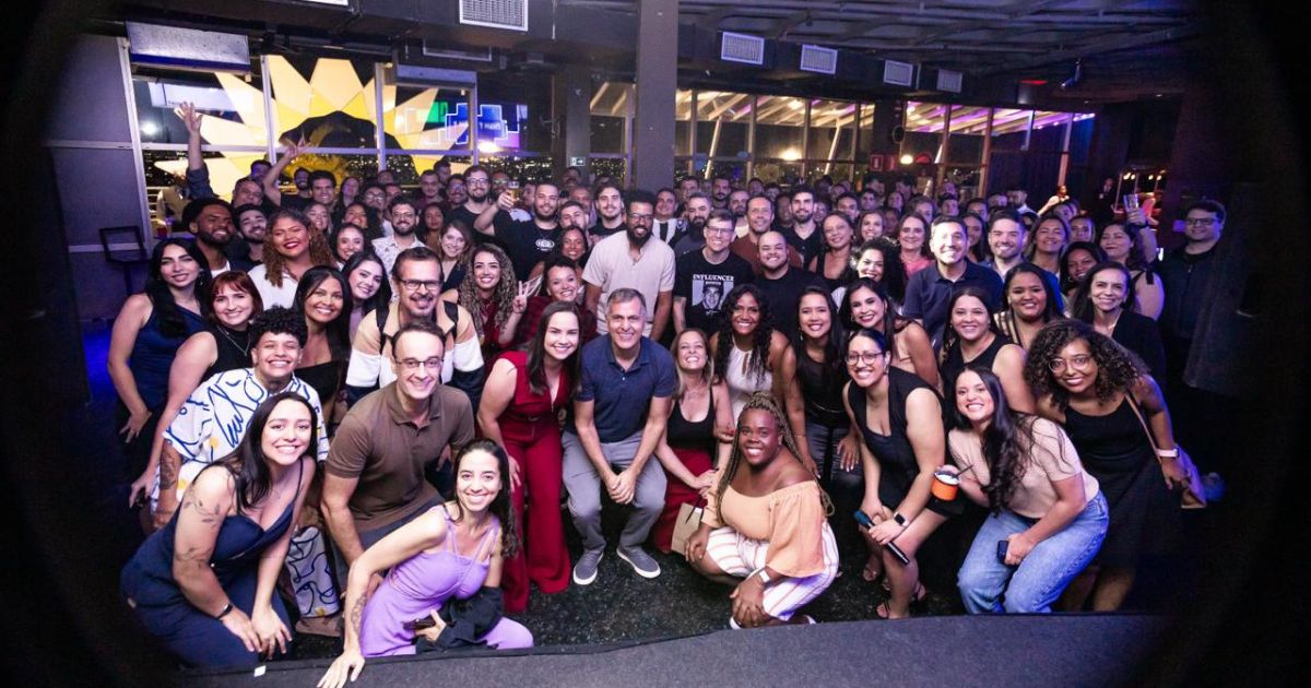 A imagem mostra um grande grupo de pessoas reunidas em um ambiente interno, que parece ser um espaço para eventos ou confraternizações. Aqui estão os principais detalhes: Configuração geral: O grupo está posicionado de frente para a câmera, ocupando praticamente todo o espaço da foto, sugerindo uma foto coletiva típica de eventos corporativos, festas ou encontros sociais. Ambiente: O local tem iluminação artificial com tons roxos e azuis, criando uma atmosfera moderna e descontraída. Ao fundo, há grandes janelas que revelam luzes externas, indicando que o evento ocorre à noite. Elementos visíveis: Estrutura do teto com dutos aparentes, reforçando um estilo industrial. Painéis iluminados e reflexos nas janelas, sugerindo um espaço sofisticado. Composição: As pessoas estão organizadas em várias fileiras, algumas agachadas na frente e outras em pé atrás, formando uma imagem de união e celebração. Essa foto transmite energia coletiva, integração e clima festivo, ideal para representar momentos de networking, comemorações ou encontros de equipe. (BHS)