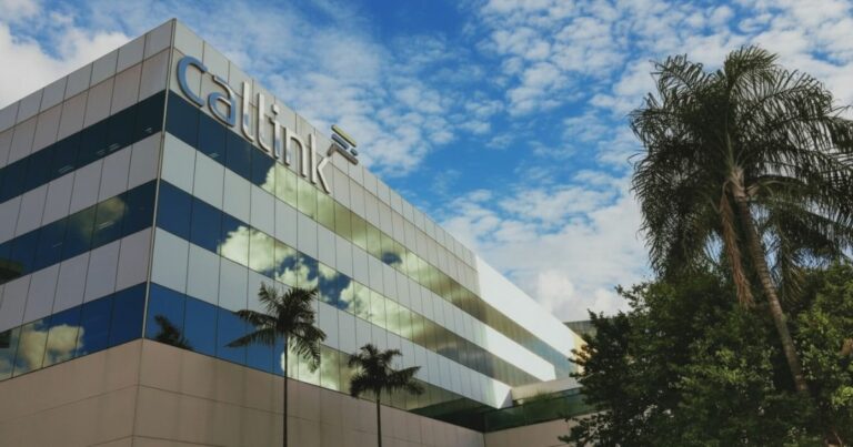 A imagem mostra a fachada de um edifício corporativo moderno com revestimento espelhado, refletindo o céu azul com nuvens brancas. Na parte superior do prédio, está visível o logotipo “Callink” em letras grandes e metálicas. Em frente ao edifício, há palmeiras e árvores, adicionando um toque natural ao cenário. A composição transmite um ambiente empresarial sofisticado, com contraste entre a arquitetura contemporânea e a vegetação.