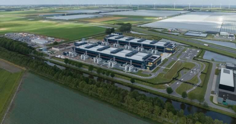 A imagem mostra uma vista aérea de um grande complexo industrial ou centro de dados, localizado em uma área rural. O complexo é composto por vários edifícios retangulares com telhados claros e estruturas externas que parecem sistemas de ventilação ou resfriamento. Ao redor do complexo, há ruas internas pavimentadas, áreas de estacionamento e canais de água que delimitam o terreno. O cenário ao fundo é formado por extensos campos verdes, estufas agrícolas e fileiras de turbinas eólicas visíveis no horizonte, indicando uma região com forte presença de agricultura e geração de energia renovável. O céu está parcialmente nublado, com nuvens brancas dispersas, e a iluminação sugere um dia claro. A imagem transmite uma sensação de infraestrutura moderna integrada a um ambiente rural. (TIP Brasil)