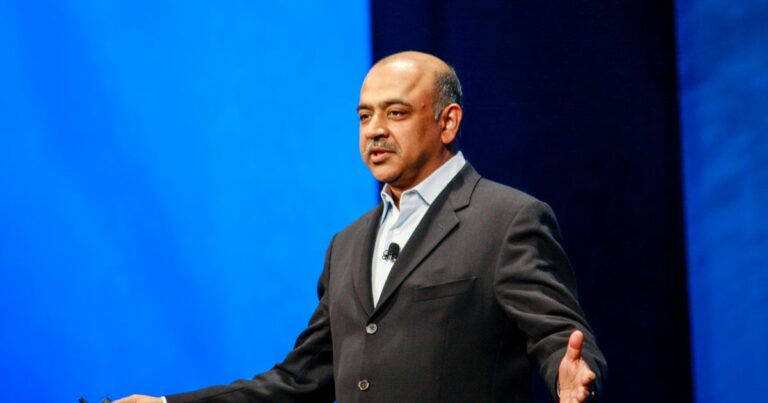 A pessoa na imagem está vestindo um terno escuro com uma camisa clara por baixo, sem gravata. Há um microfone preso à lapela, indicando que provavelmente está participando de uma apresentação ou palestra. O fundo é composto por um grande painel azul, sugerindo um ambiente de conferência ou evento corporativo. A postura, com braços abertos e mãos visíveis, transmite uma atitude de explicação ou engajamento com o público. A iluminação é forte e direcionada, típica de palcos ou auditórios. (AGI)