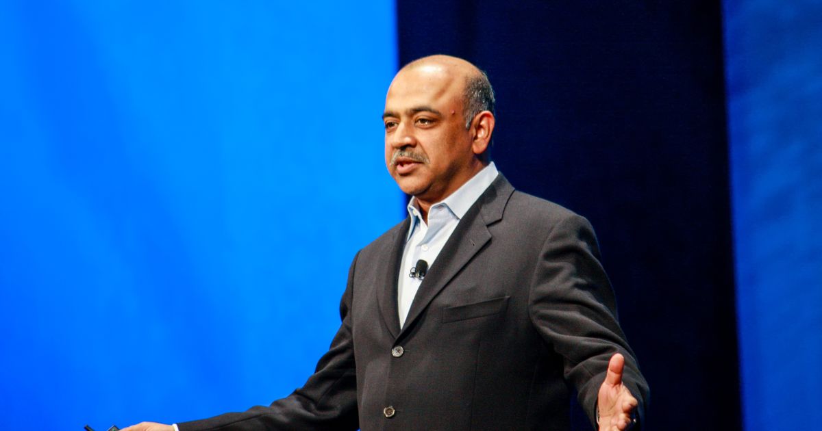 A pessoa na imagem está vestindo um terno escuro com uma camisa clara por baixo, sem gravata. Há um microfone preso à lapela, indicando que provavelmente está participando de uma apresentação ou palestra. O fundo é composto por um grande painel azul, sugerindo um ambiente de conferência ou evento corporativo. A postura, com braços abertos e mãos visíveis, transmite uma atitude de explicação ou engajamento com o público. A iluminação é forte e direcionada, típica de palcos ou auditórios. (AGI)