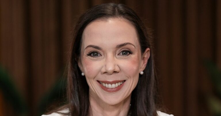 A imagem mostra a parte superior de uma mulher com cabelos longos e escuros, penteados para trás. Ela usa brincos de pérola e está vestida com uma roupa clara. O fundo apresenta um ambiente interno desfocado, com tons terrosos e elementos verdes que parecem ser plantas, criando uma atmosfera elegante e acolhedora. (Cíntia Scovine Barcelos, CTO do Bradesco)