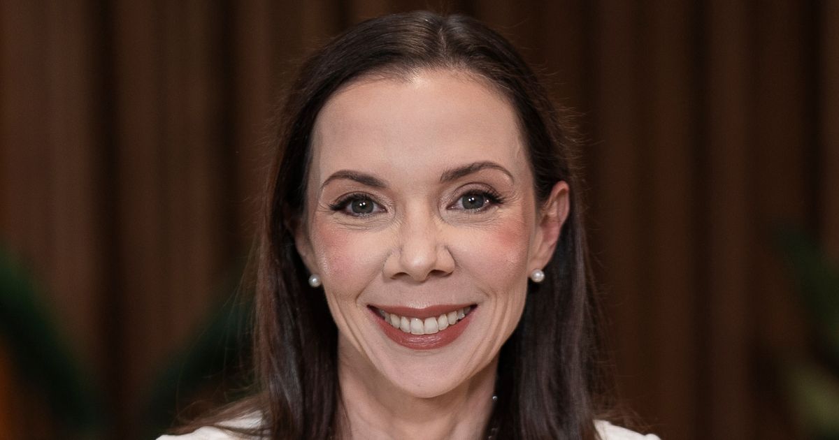 A imagem mostra a parte superior de uma mulher com cabelos longos e escuros, penteados para trás. Ela usa brincos de pérola e está vestida com uma roupa clara. O fundo apresenta um ambiente interno desfocado, com tons terrosos e elementos verdes que parecem ser plantas, criando uma atmosfera elegante e acolhedora. (Cíntia Scovine Barcelos, CTO do Bradesco)