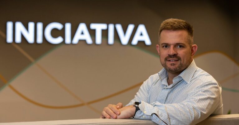 Um homem está sentado apoiando os antebraços sobre uma superfície clara, em um ambiente interno moderno. Ele veste uma camisa social quadriculada em tons claros e um relógio escuro no pulso esquerdo. Ao fundo, há uma parede com iluminação suave e o texto “INICIATIVA” em destaque, acompanhado por linhas curvas decorativas em cores discretas que compõem um visual corporativo e contemporâneo. (Afranio Lamy Spolador Junior, diretor de Tecnologia e Inovação da Ecorodovias)