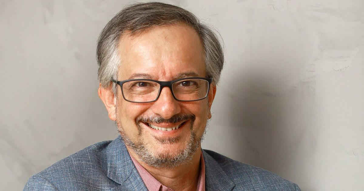 A imagem mostra a parte superior de um homem posicionado diante de uma parede de textura clara. Ele veste um blazer azul acinzentado sobre uma camisa social rosa, compondo um visual formal e discreto. O enquadramento destaca a roupa e os ombros, com iluminação suave que cria um tom neutro e profissional. (Nelson Campelo, CEO da Atos para a América do Sul)