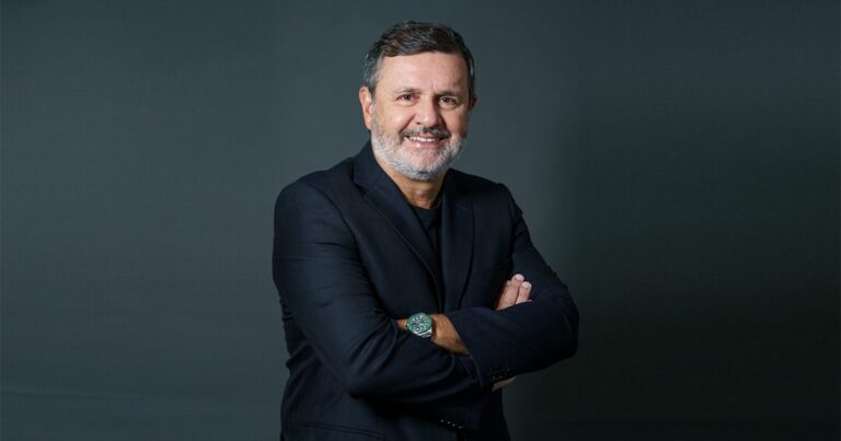 Um homem está posando diante de um fundo escuro e uniforme, usando um terno preto com camiseta preta por baixo. Ele mantém os braços cruzados, exibindo um relógio metálico no pulso esquerdo. A postura transmite confiança e profissionalismo, com iluminação suave destacando o contraste entre a roupa e o fundo. (Vittorio Danesi, CEO da Simpress)