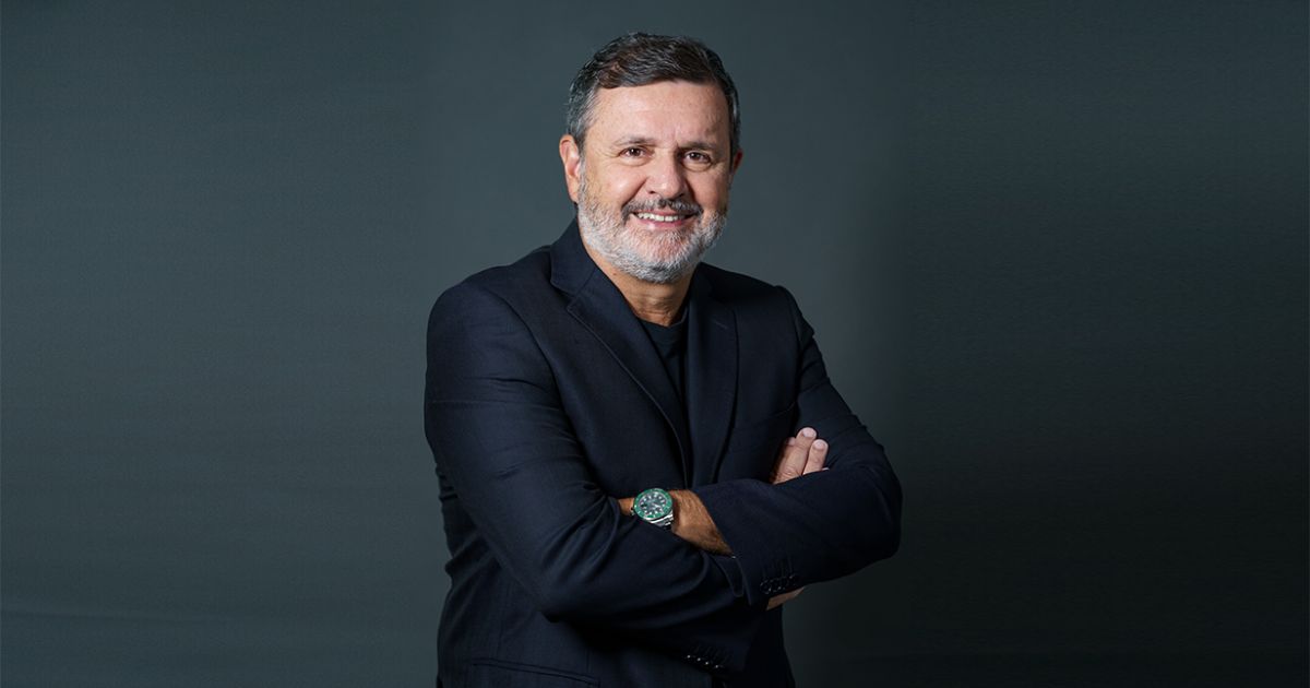 Um homem está posando diante de um fundo escuro e uniforme, usando um terno preto com camiseta preta por baixo. Ele mantém os braços cruzados, exibindo um relógio metálico no pulso esquerdo. A postura transmite confiança e profissionalismo, com iluminação suave destacando o contraste entre a roupa e o fundo. (Vittorio Danesi, CEO da Simpress)