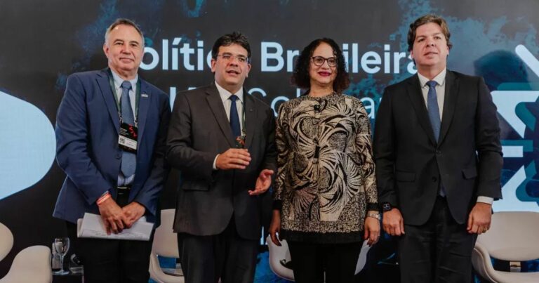 A imagem mostra quatro pessoas em pé, lado a lado, em um ambiente formal, provavelmente um evento ou conferência. Todas estão vestidas com roupas sociais: três delas usam ternos escuros com gravata, enquanto a pessoa ao centro veste uma blusa estampada em tons de preto, branco e bege, com padrões abstratos. Ao fundo, há um painel com texto parcialmente visível, incluindo as palavras “Política Brasileira” e outros caracteres que parecem fazer parte de um título ou tema do evento. Também é possível ver cadeiras brancas e copos sobre uma mesa baixa, sugerindo um espaço preparado para palestras ou debates. A iluminação é clara e destaca os participantes contra o fundo escuro com elementos gráficos. (infraestrutura)
